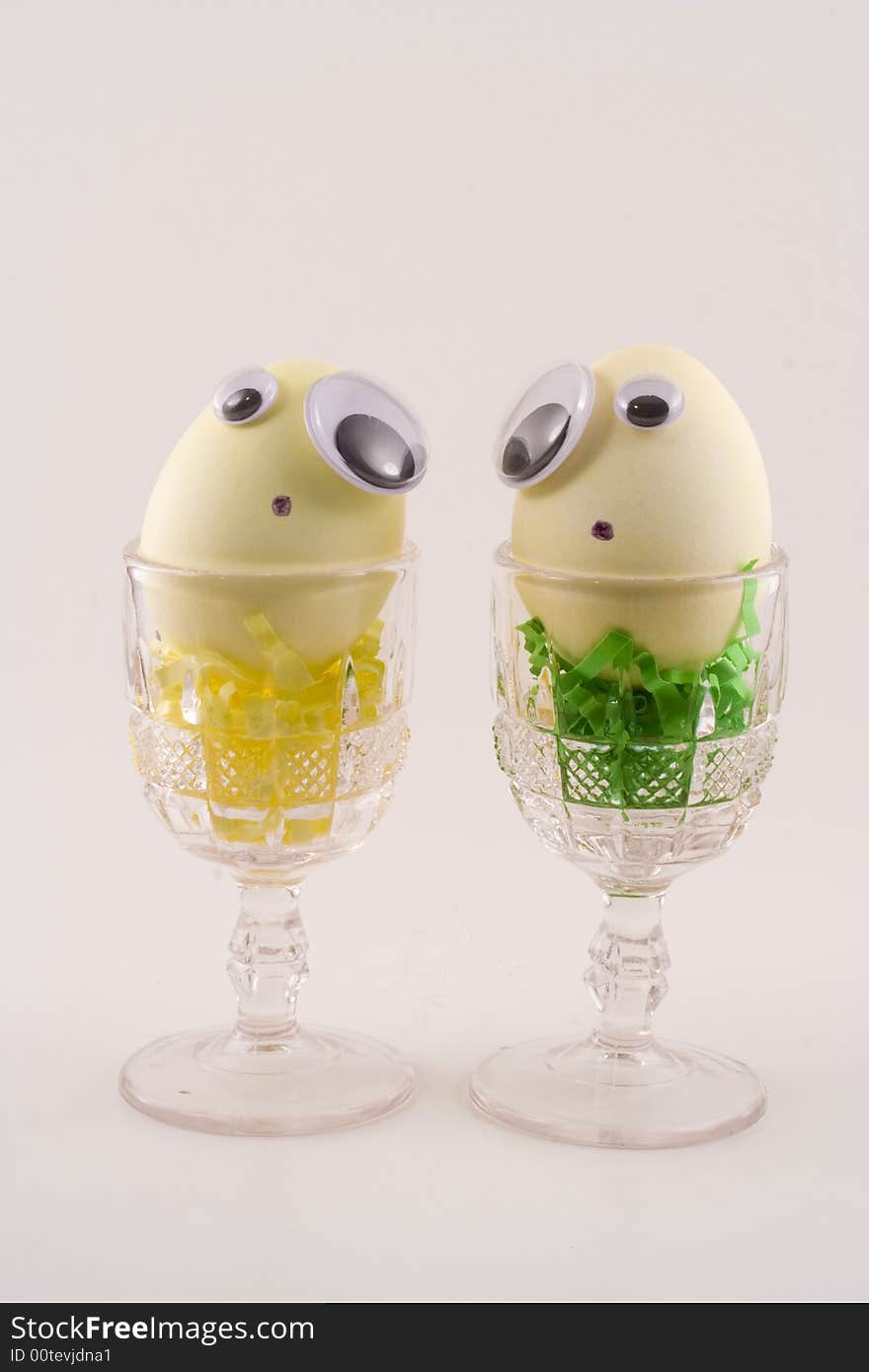 Two yellow Easter eggs with googly eyes sit in cups and make faces at each other. Two yellow Easter eggs with googly eyes sit in cups and make faces at each other.