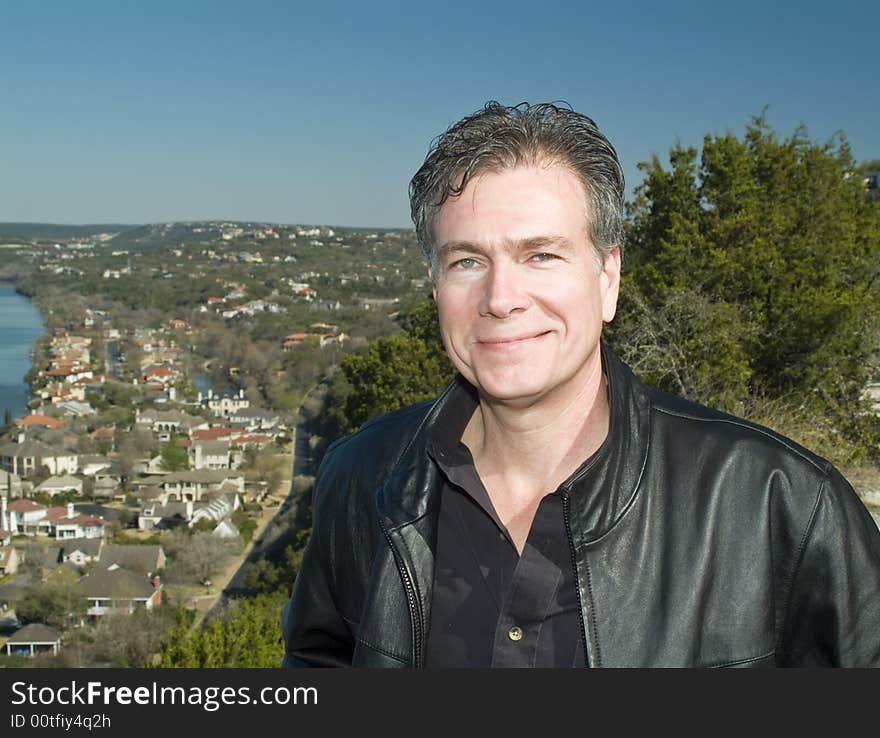 A smiling man on a hilltop with an affluential riverside neighborhood in the valley below him. A smiling man on a hilltop with an affluential riverside neighborhood in the valley below him.