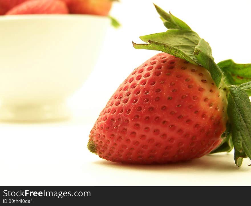 Close up image of a large red strawberry, with other strawberries in a bowl in the background.  White background and horizontal orientation. Close up image of a large red strawberry, with other strawberries in a bowl in the background.  White background and horizontal orientation.