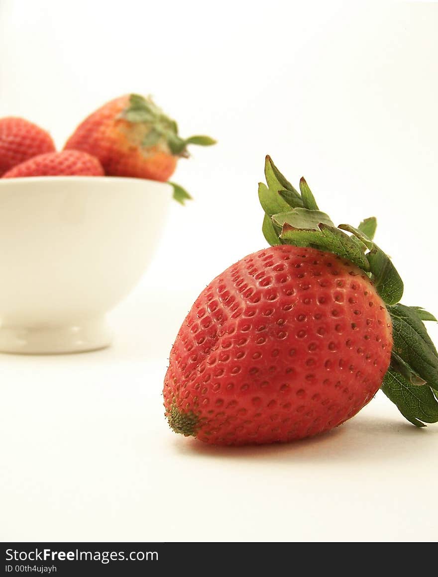 Image of a bright red strawberry in the foreground, and a small white bowl with other strawberries in the background.  White background.  Vertical orientation. Image of a bright red strawberry in the foreground, and a small white bowl with other strawberries in the background.  White background.  Vertical orientation.