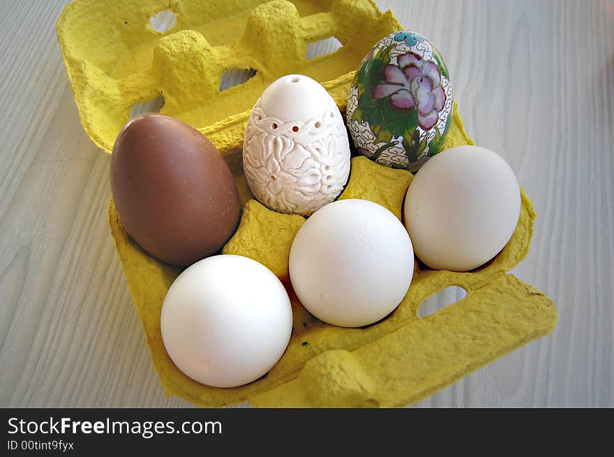 A chocolate egg, a chinese decorated porcelain egg, a hand carved meerschaum egg and three white eggs in a yellow egg box on white and gray oak laminate. A chocolate egg, a chinese decorated porcelain egg, a hand carved meerschaum egg and three white eggs in a yellow egg box on white and gray oak laminate