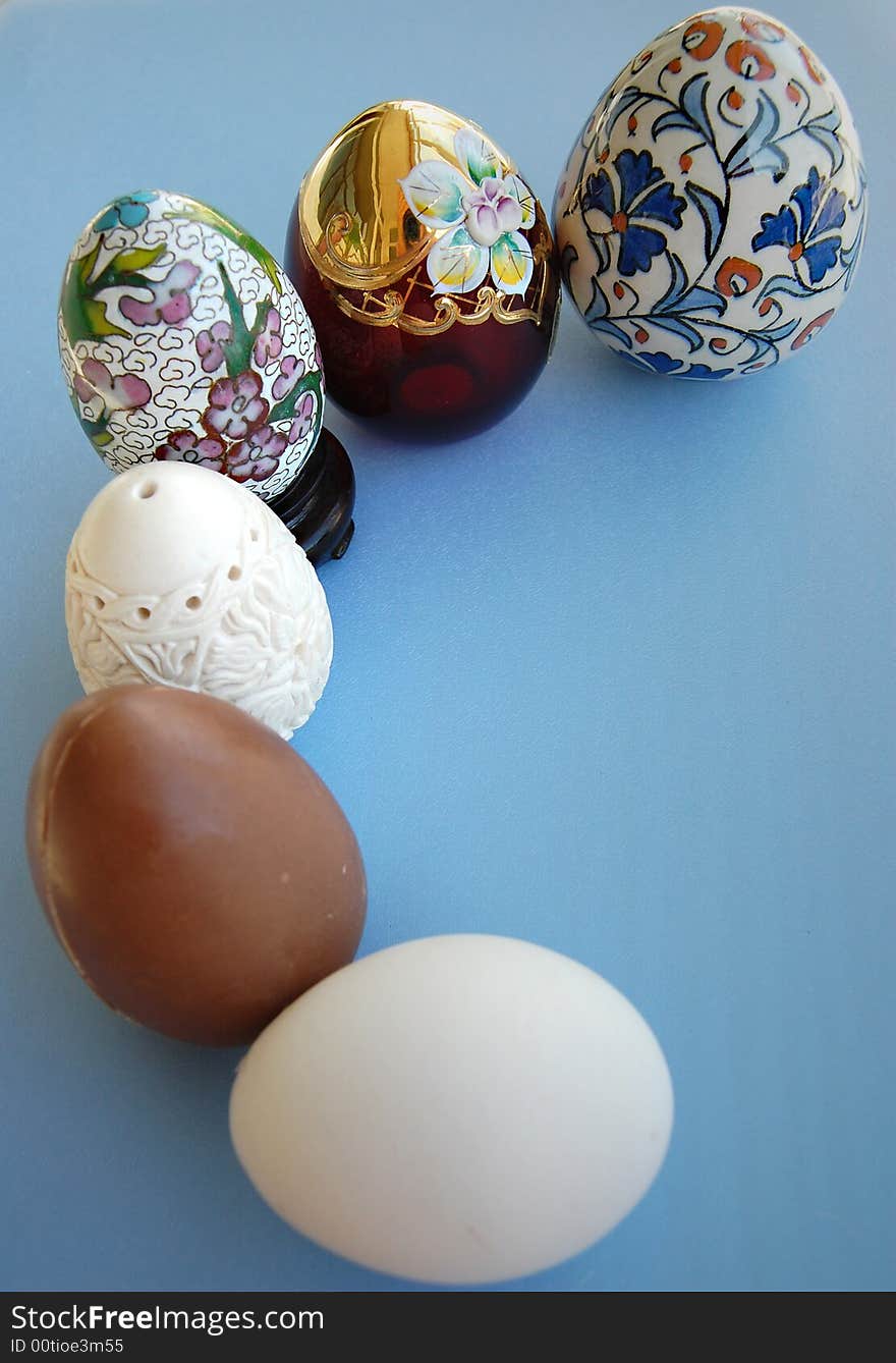 A chocolate egg, a Chinese decorated porcelain egg, a hand carved meerschaum egg, a burgundy and gold glass egg and a white eggs on blue background together. A chocolate egg, a Chinese decorated porcelain egg, a hand carved meerschaum egg, a burgundy and gold glass egg and a white eggs on blue background together