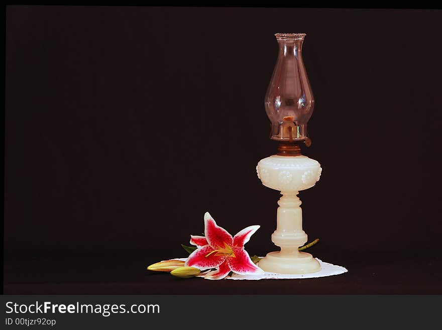 A milk glass oil lanp from the late 1800s. It has a decorative around the bowl, mantle and wick turner. Ared lily complements the composition. A milk glass oil lanp from the late 1800s. It has a decorative around the bowl, mantle and wick turner. Ared lily complements the composition