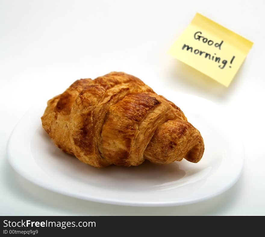 A fresh croissant in a white plate and a yellow note with this text: 'Good morning!'. A fresh croissant in a white plate and a yellow note with this text: 'Good morning!'