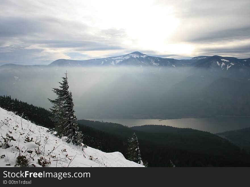 A lone wind swept tree stands in the snow overlooking a river with mountains in the background. A lone wind swept tree stands in the snow overlooking a river with mountains in the background