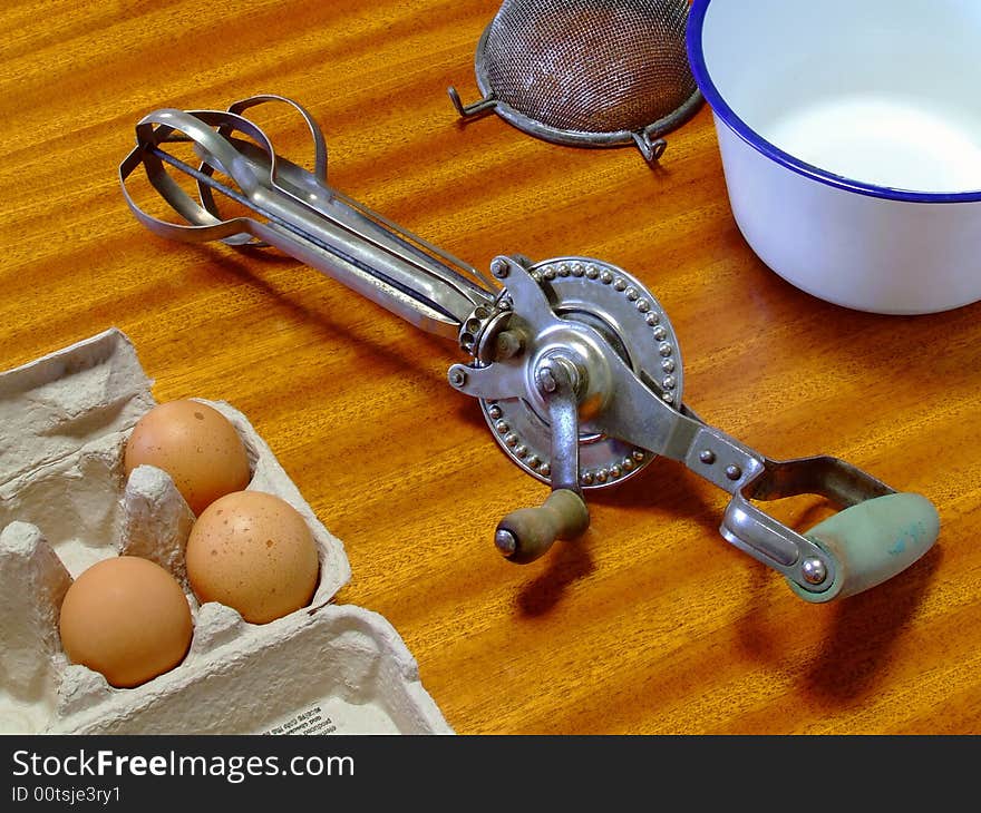 An old and well-used manual egg-beater lying on a table, with related items. An old and well-used manual egg-beater lying on a table, with related items.