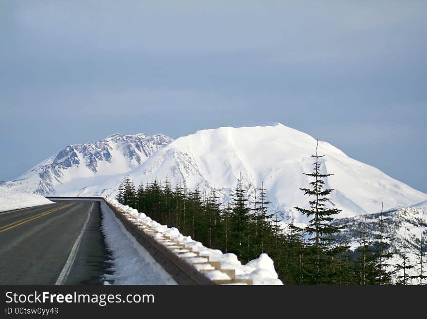 Paved road with snow packed guardrail, the peak of snow covered mountain, blue sky, evergreen trees in foreground. Paved road with snow packed guardrail, the peak of snow covered mountain, blue sky, evergreen trees in foreground