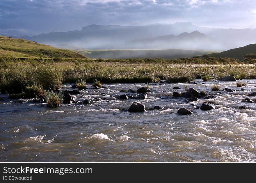 Flowing water of Tugela River with the Amphitheatre at the background in the evening light. Northern Drakensberg, South Africa. Flowing water of Tugela River with the Amphitheatre at the background in the evening light. Northern Drakensberg, South Africa.