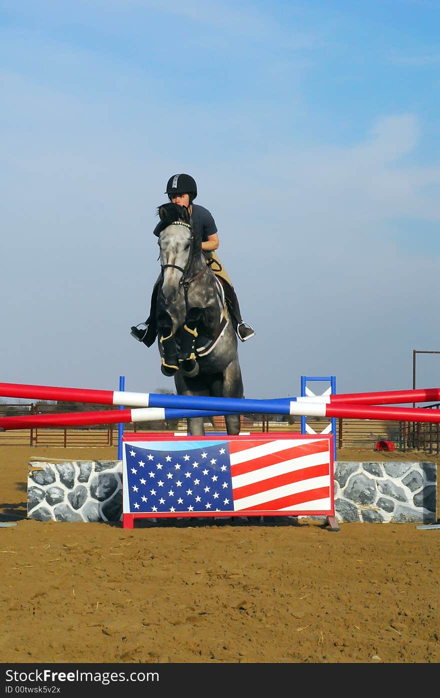 Young woman riding dappled grey horse over jump with painted Amercian flag. Young woman riding dappled grey horse over jump with painted Amercian flag.