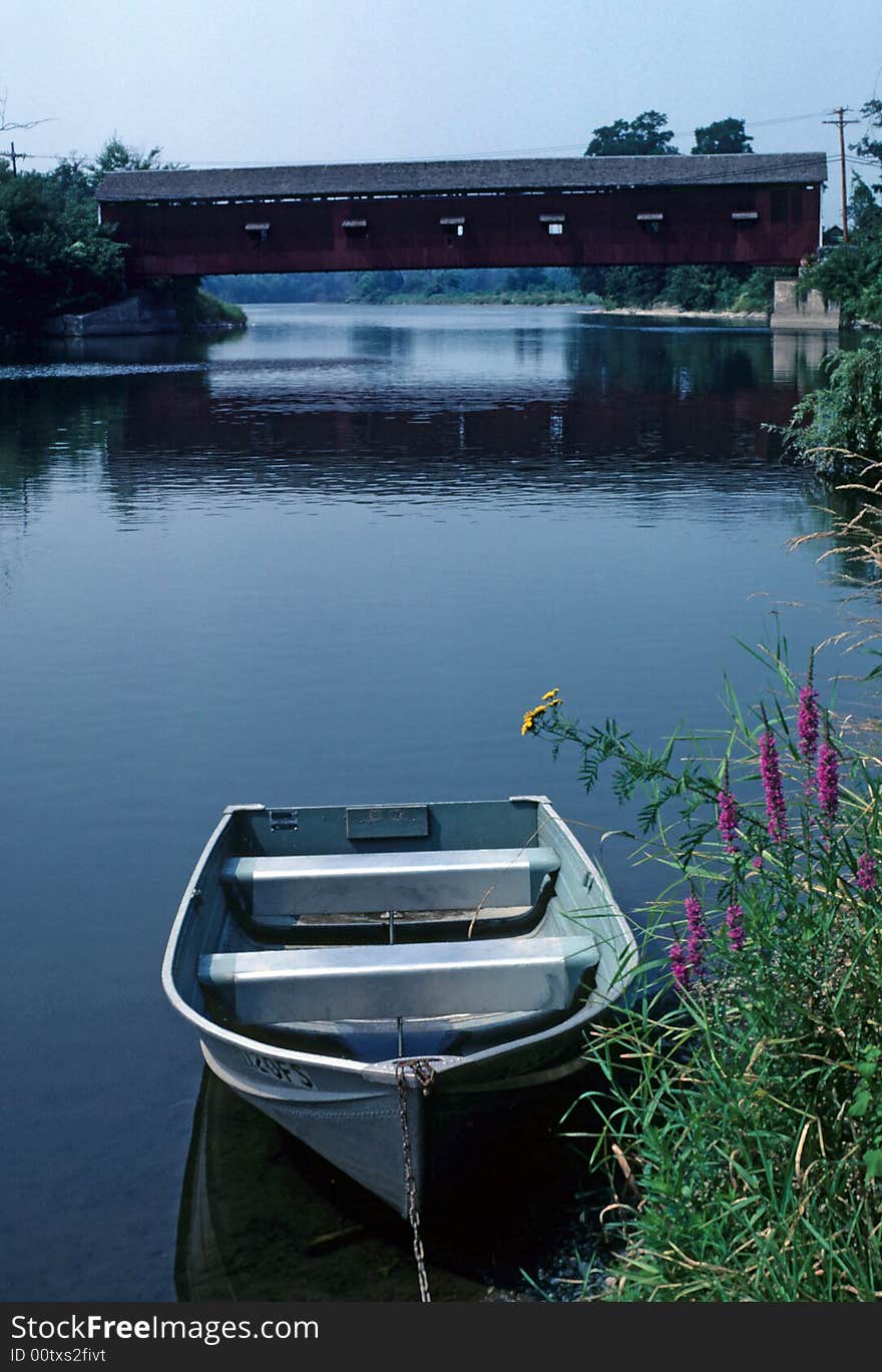 A row boat sits tied to the shore with a covered bridge in the background