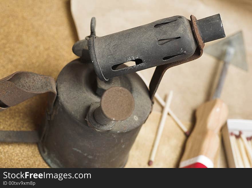 An old blow-lamp with glass-paper, matches and a paint scraper in the background.