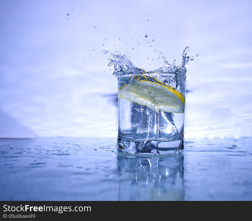 Glass of cold clear splashing water with falling lemon's slice on light-blue background. Glass of cold clear splashing water with falling lemon's slice on light-blue background