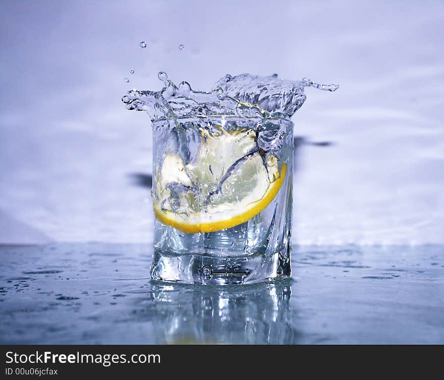 Glass of cold clear splashing water with falling lemon's slice on light-blue background. Glass of cold clear splashing water with falling lemon's slice on light-blue background