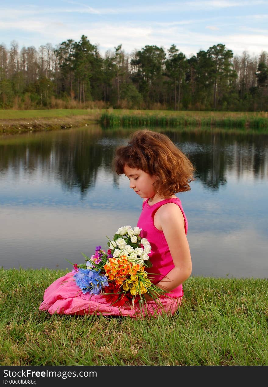 A pretty little girl is sitting on the grass holding flowers on the lake background. A pretty little girl is sitting on the grass holding flowers on the lake background