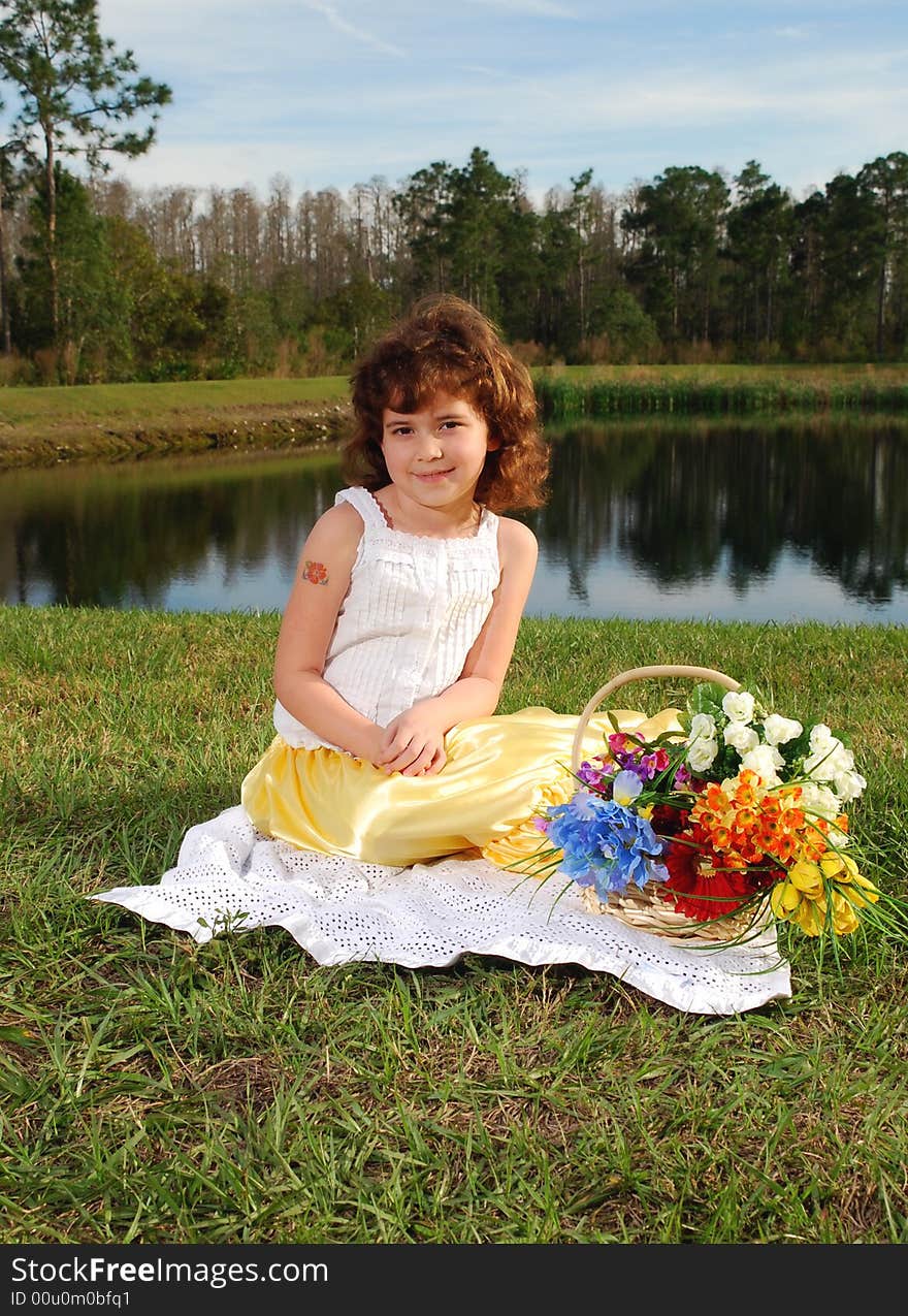 A pretty little  girl is sitting on the grass with the basket of flowers next to her on the lake background. A pretty little  girl is sitting on the grass with the basket of flowers next to her on the lake background