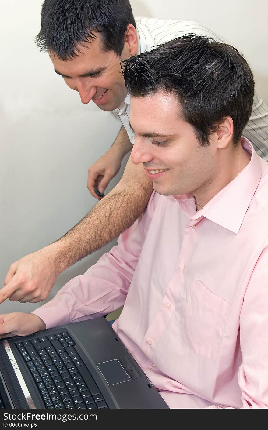 Two young men, working on a laptop computer, one sitting in his chair, and the other, standing next to him, both smiling. Two young men, working on a laptop computer, one sitting in his chair, and the other, standing next to him, both smiling