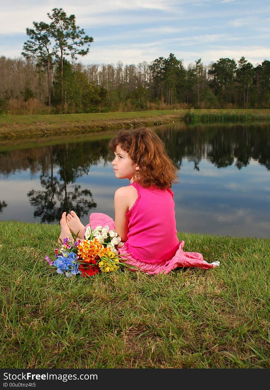 A cute girl in pink outfit is sitting at the lake with flowers bouquet next to her. A cute girl in pink outfit is sitting at the lake with flowers bouquet next to her