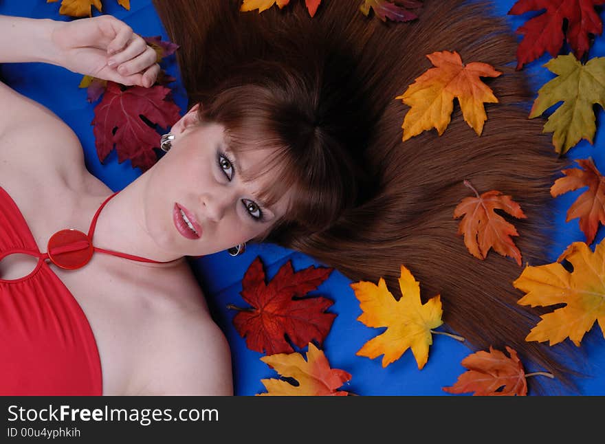 Cute brunette girl with hair spread around, laying on blue background, surrounded by maple leaves. Cute brunette girl with hair spread around, laying on blue background, surrounded by maple leaves