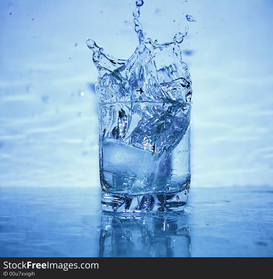 Glass of splashing water with ice on blue background