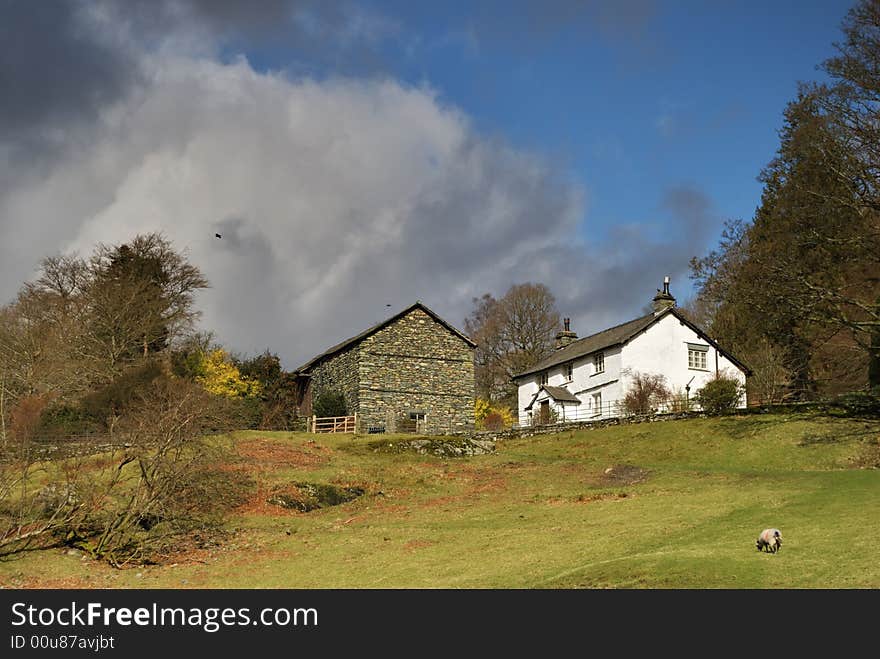 A White farmhouse and stone barn standing together on a sunny slope with dark clouds