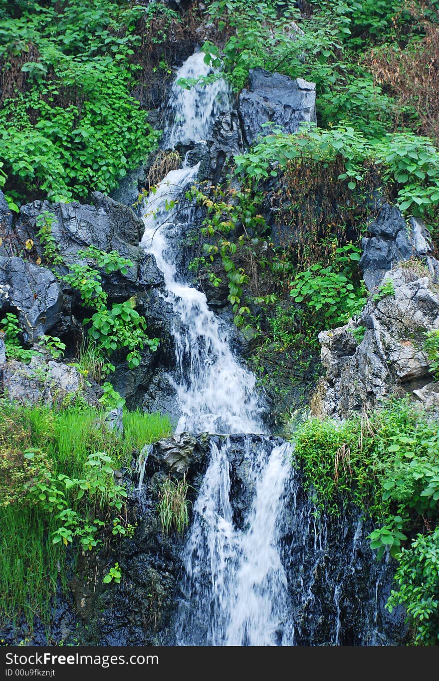 A little cascde at the foot of a stone hill,pure and clear water flows down with flourish vegetation. A little cascde at the foot of a stone hill,pure and clear water flows down with flourish vegetation.