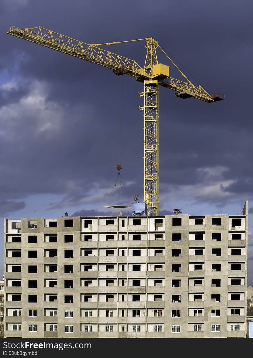 Hoisting crane lifting a concrete slab on a house being built, on the background of gloomy thunder clouds. Hoisting crane lifting a concrete slab on a house being built, on the background of gloomy thunder clouds