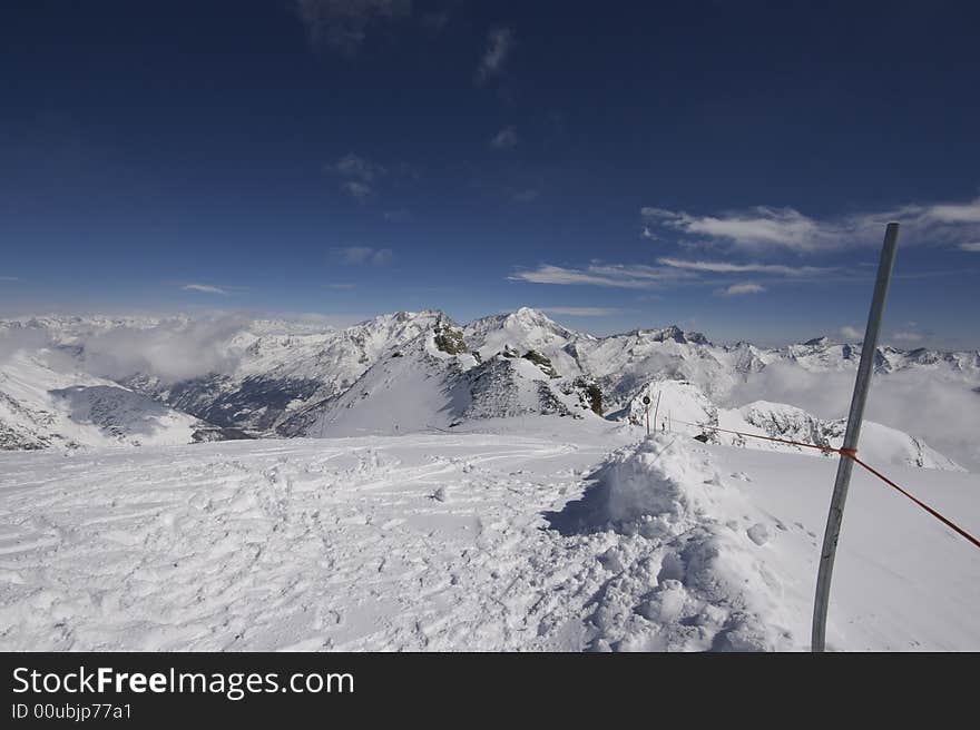 This photograph shows the top of a mountain in the resort of Saas-Fee in Switzerland. There are ski marks in the snow, and you can see down the valley over Saas-Fee and other mountain villages. This photograph shows the top of a mountain in the resort of Saas-Fee in Switzerland. There are ski marks in the snow, and you can see down the valley over Saas-Fee and other mountain villages.