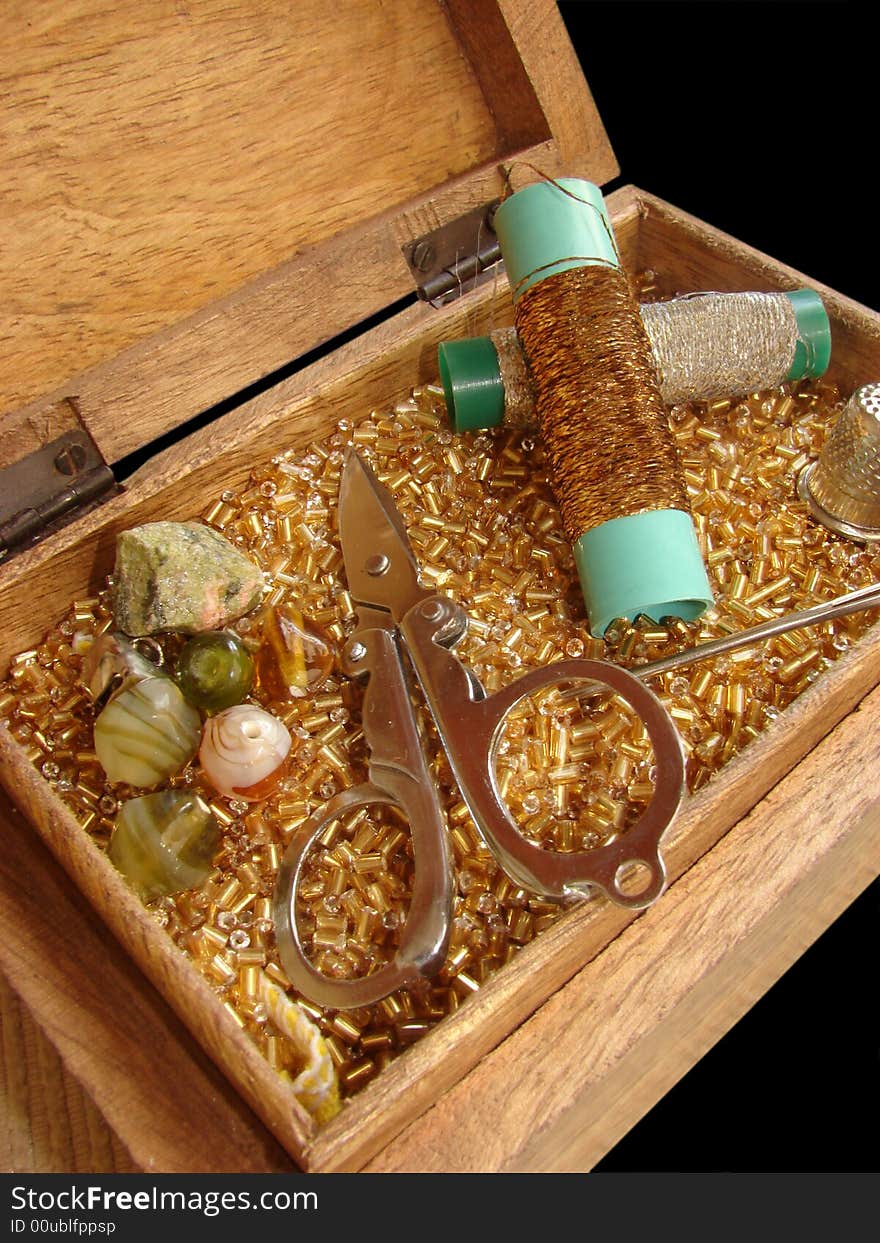 Sewing basket in golden colors, containing scissors, beads, stones, golden and silver thread, thimble and needle on a black background.