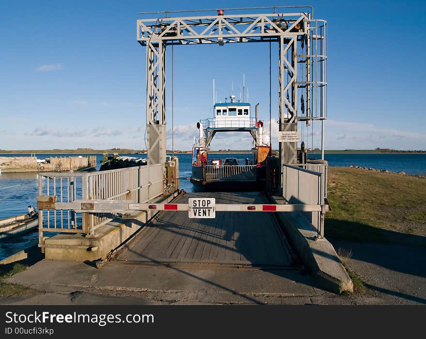 A black and white ferry leaving a small terminal. The road is blocked by a boom with a stop sign. A black and white ferry leaving a small terminal. The road is blocked by a boom with a stop sign.