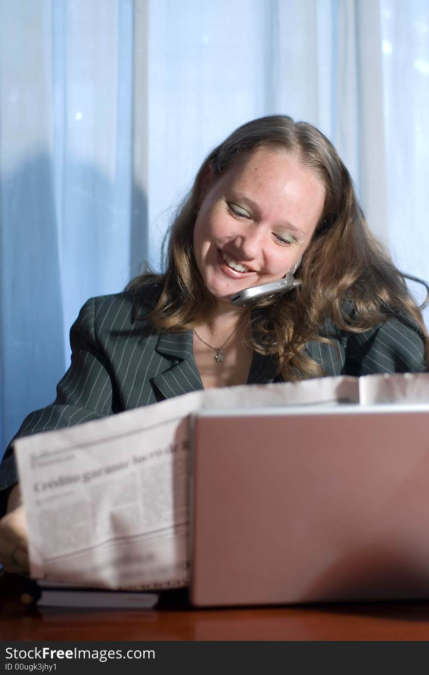 A woman, talking on a cellphone, sitting holding a newspaper behind a laptop. A woman, talking on a cellphone, sitting holding a newspaper behind a laptop.