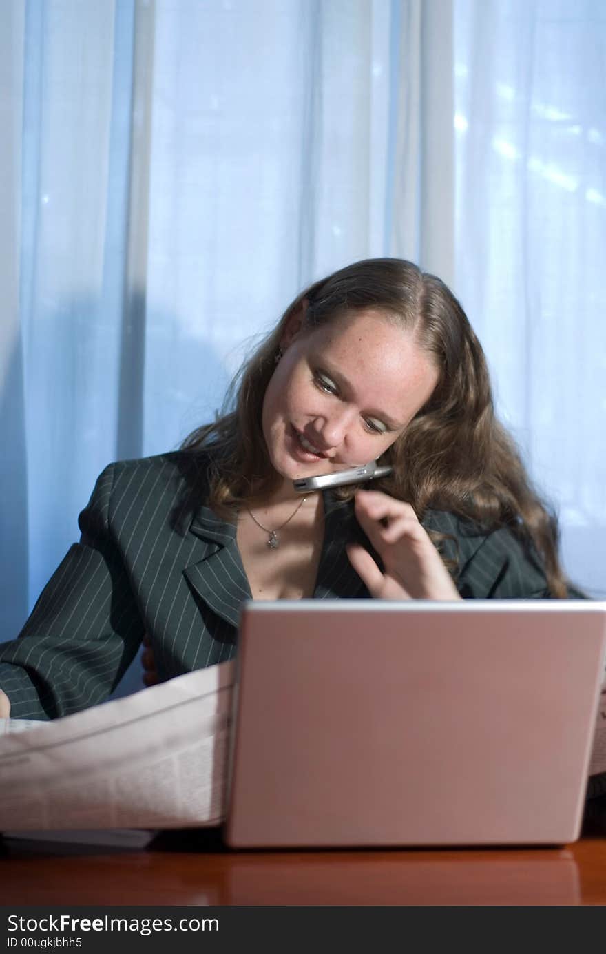 A woman, talking on a cellphone, sitting holding a newspaper behind a laptop. A woman, talking on a cellphone, sitting holding a newspaper behind a laptop.