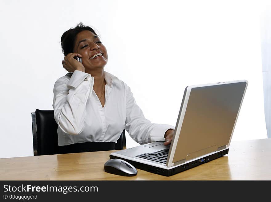 Indian business woman leaning back and laughing while on her phone sitting in front of her laptop. Indian business woman leaning back and laughing while on her phone sitting in front of her laptop