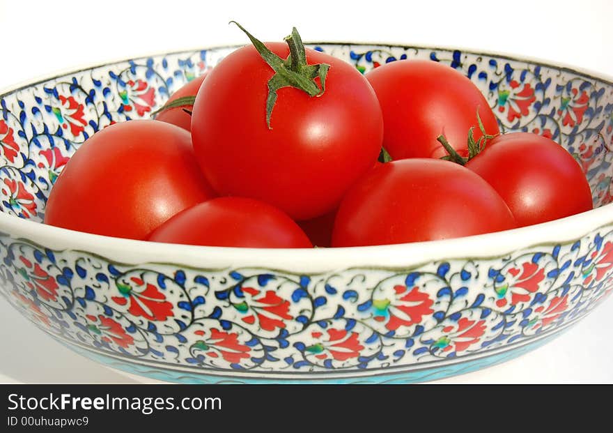 Glossy red ripe round small tomatos with stalks in a hand painted Iznik ceramic bowl with classical red, green, blue, turquoise carnation patterns on white. Glossy red ripe round small tomatos with stalks in a hand painted Iznik ceramic bowl with classical red, green, blue, turquoise carnation patterns on white.