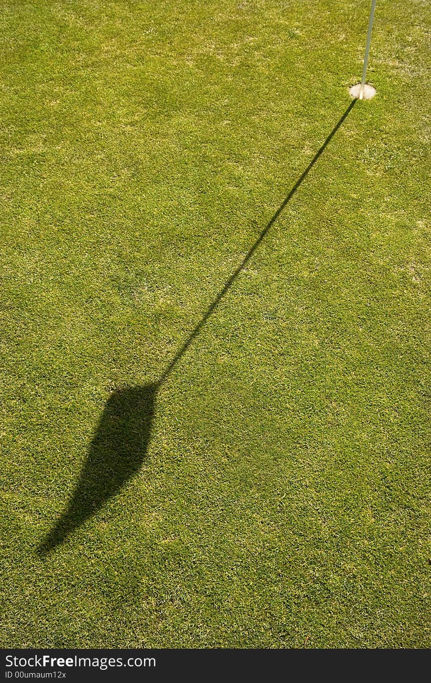 On the Putting green of a golf course the flag and pin or stick casting a shadow on the green. On the Putting green of a golf course the flag and pin or stick casting a shadow on the green