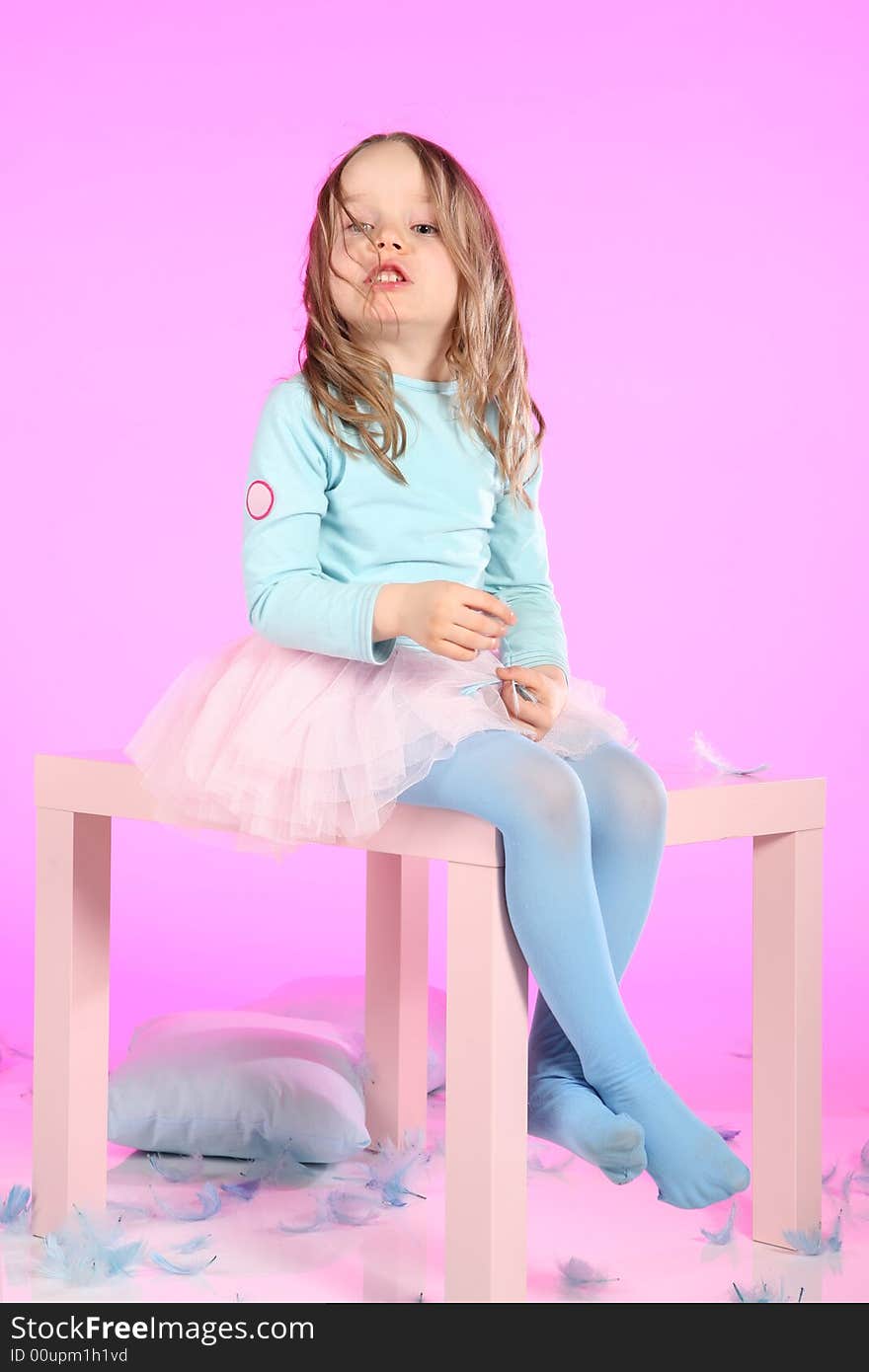 Five years old girl having fun with blue pillows and feathers at small table on pink colored background. Studio hi-res shot. Five years old girl having fun with blue pillows and feathers at small table on pink colored background. Studio hi-res shot.