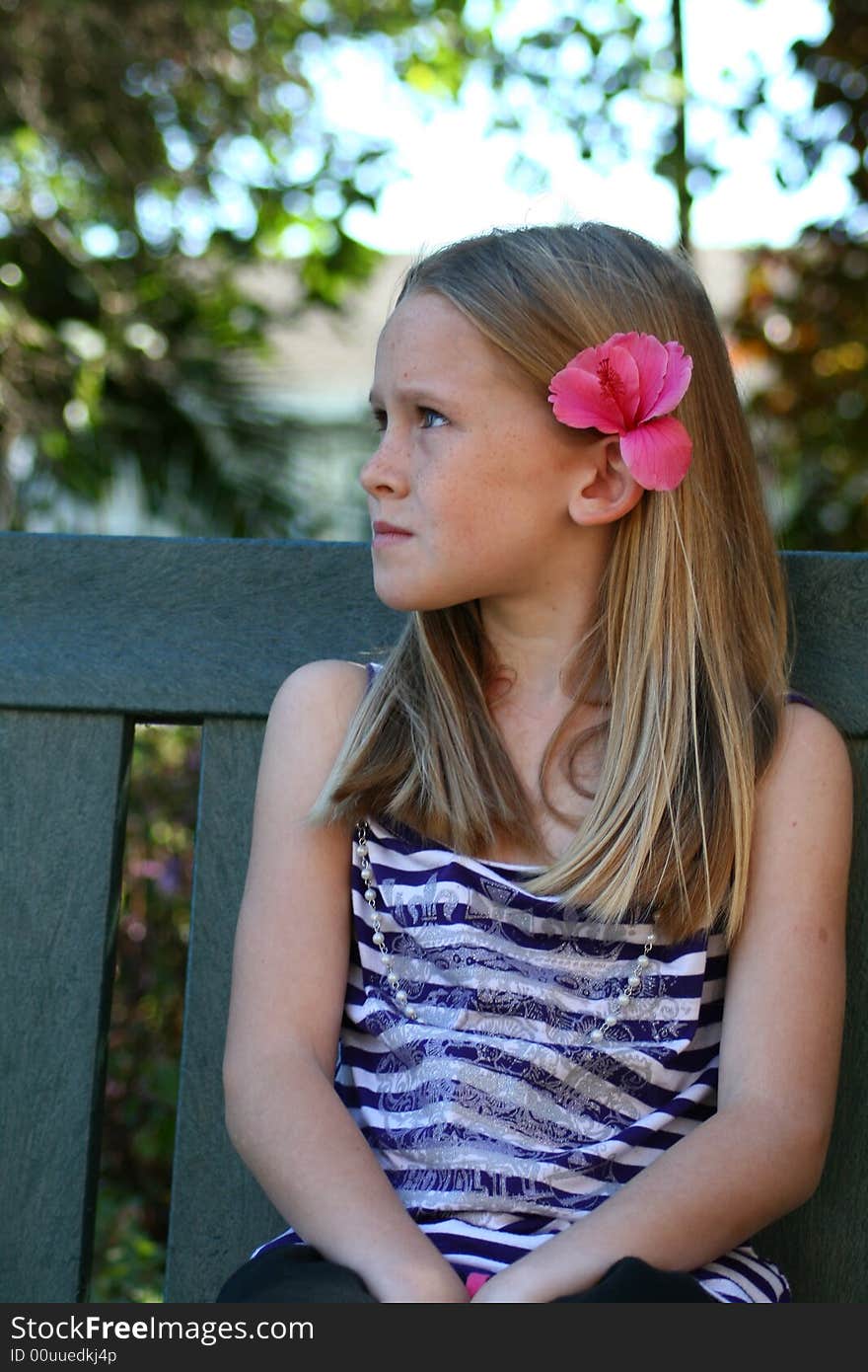 A white caucasian girl sitting on a bench in the garden with a bored expression on her face with a pink flower in her hair. A white caucasian girl sitting on a bench in the garden with a bored expression on her face with a pink flower in her hair