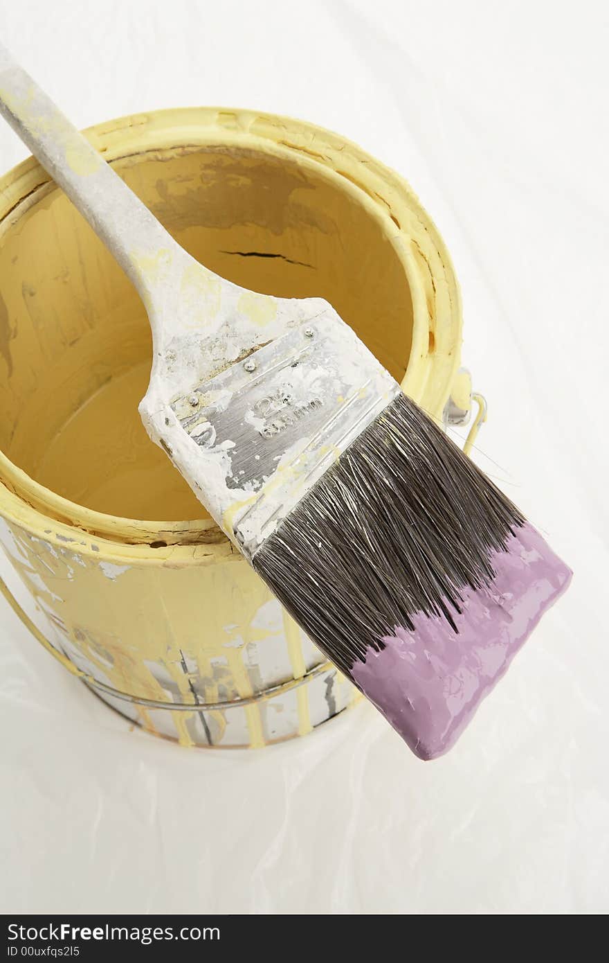 A metallic paint can on top off a plastic drop cloth. The paint can has yellow paint inside. The brush is dipped in purple paint. A metallic paint can on top off a plastic drop cloth. The paint can has yellow paint inside. The brush is dipped in purple paint.