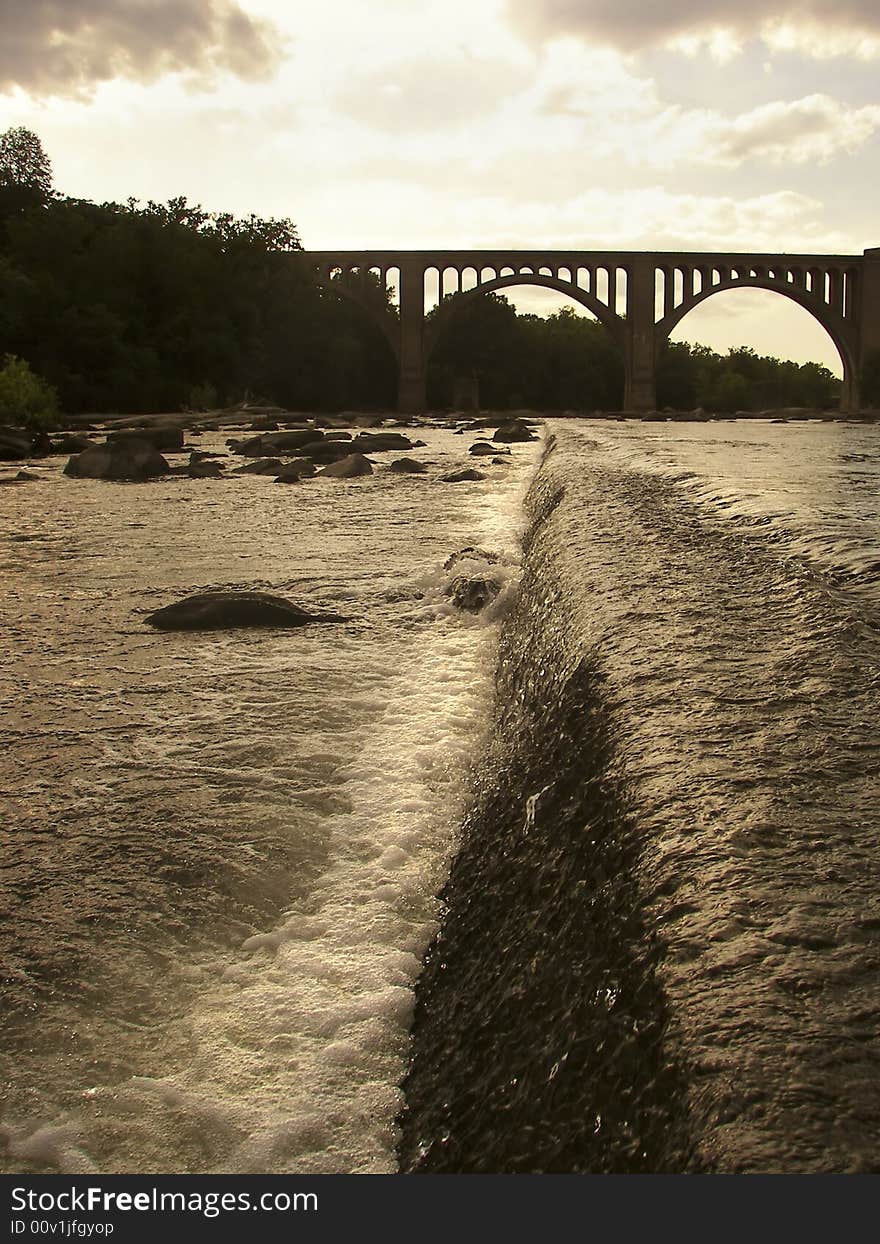 View down a dam looking at a bridge that is crossing the river in the distance. View down a dam looking at a bridge that is crossing the river in the distance.