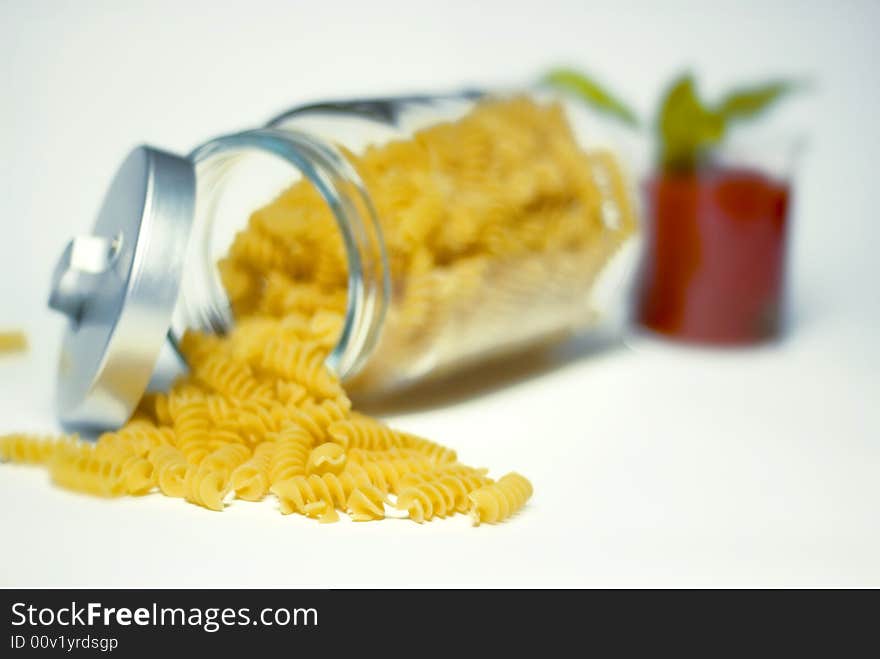 Italian fusilli pasta falling out of a glass jar with a bowl of tomato sauce and basil on the back ground. Italian fusilli pasta falling out of a glass jar with a bowl of tomato sauce and basil on the back ground