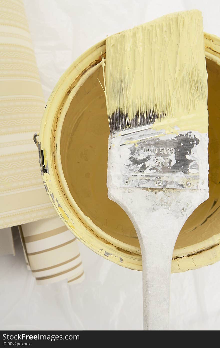 Brush dipped in yellow paint on top of a can of paint with wall paper samples on the side. Brush dipped in yellow paint on top of a can of paint with wall paper samples on the side.