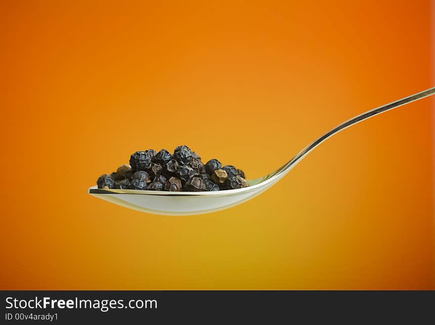 Metal spoon filled with black peppers over orange background