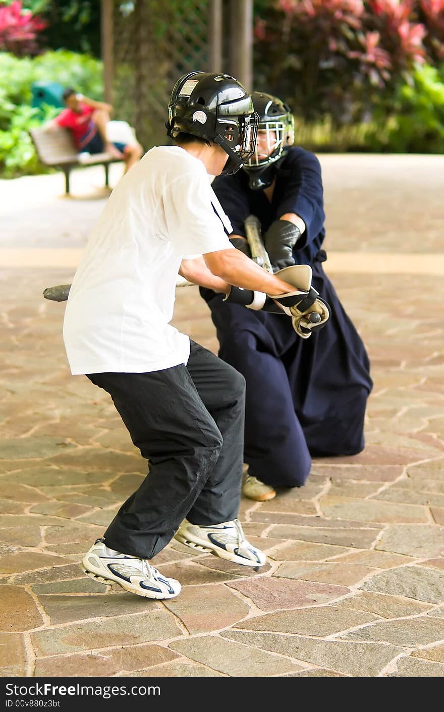 Free weapon fighting enthusiasts, a lance versus a sword and shield, sparring in an open shaded public space on a weekend. Free weapon fighting enthusiasts, a lance versus a sword and shield, sparring in an open shaded public space on a weekend