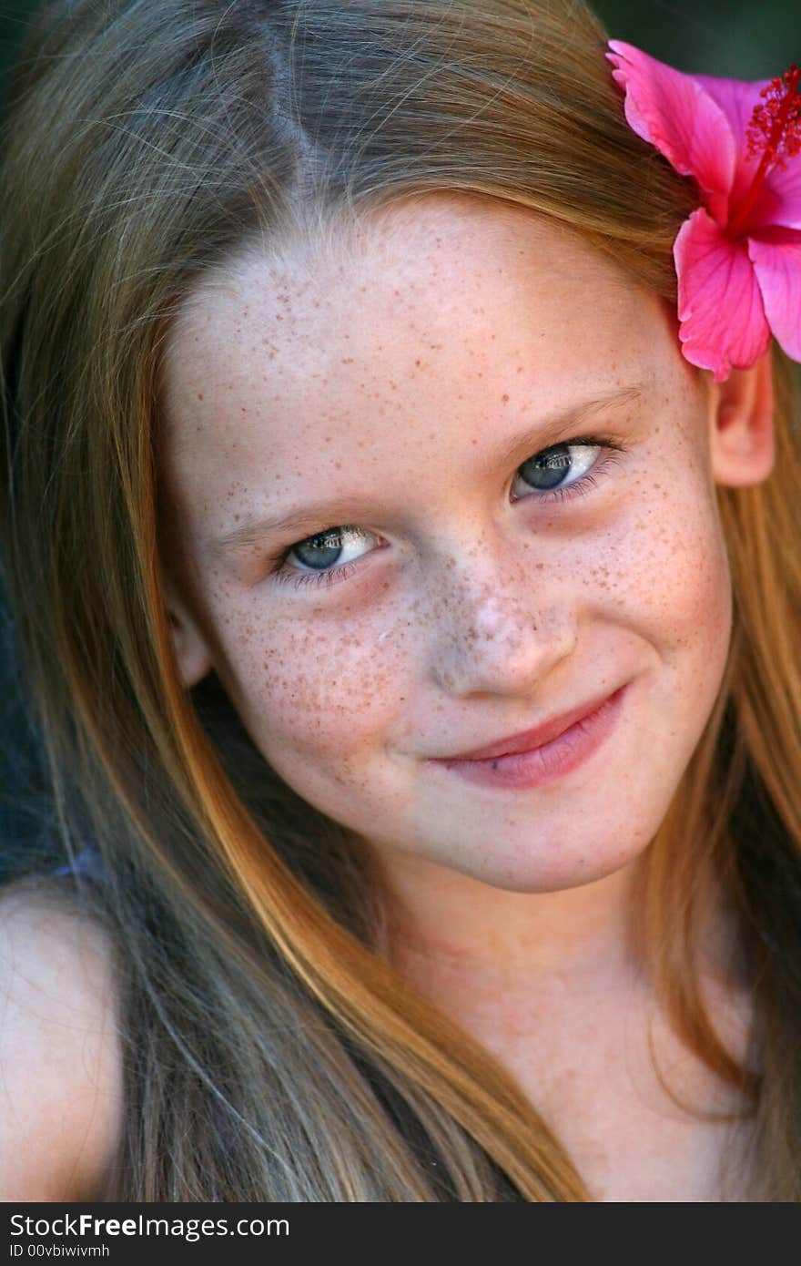 A white caucasian girl child with a pink flower in her hair smiling softly. A white caucasian girl child with a pink flower in her hair smiling softly