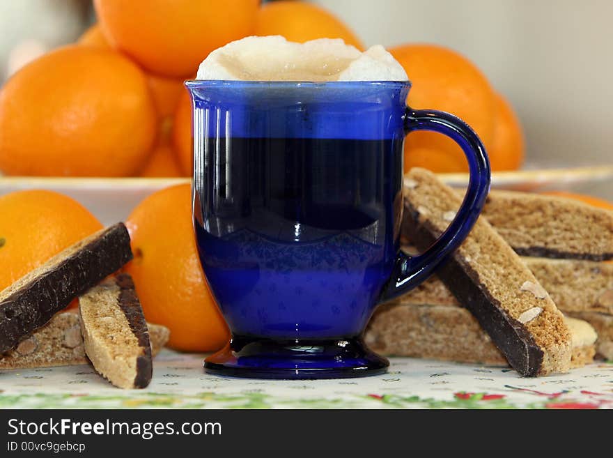 Coffee latte in cobalt blue glass mug surrounded by chocolate biscotti and fresh oranges. Coffee latte in cobalt blue glass mug surrounded by chocolate biscotti and fresh oranges