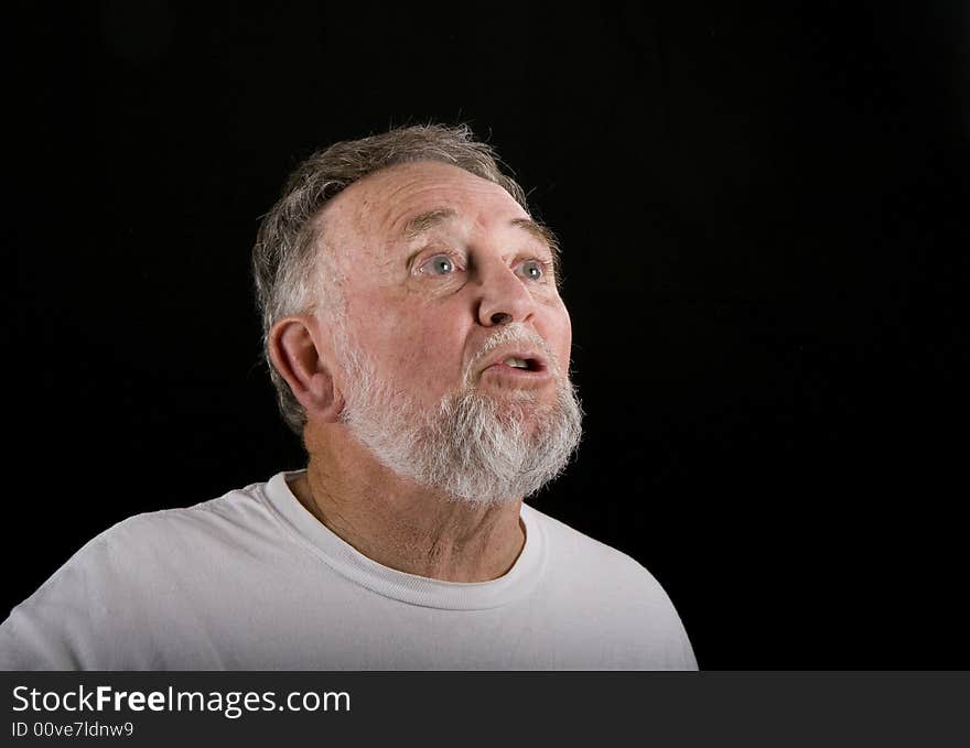 An older man in a white t-shirt on a black background looking up into copy space with shocked expression. An older man in a white t-shirt on a black background looking up into copy space with shocked expression