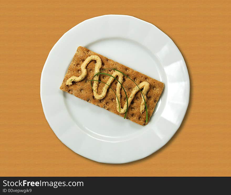 A view of a large multi-grained cracker on a plate or saucer and topped with a wavy line of fish spread and green garnish. A view of a large multi-grained cracker on a plate or saucer and topped with a wavy line of fish spread and green garnish.