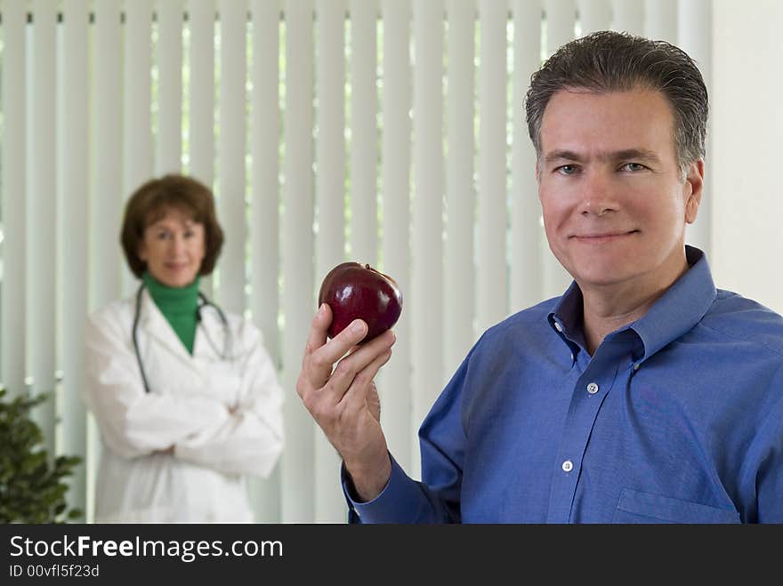 A man with an apple in his hand with a smiling woman dressed as a doctor, out of focus in the background. A man with an apple in his hand with a smiling woman dressed as a doctor, out of focus in the background.