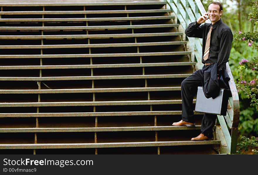 A businessman in a black pinstripe suit, holding a briefcase, standing at the base of outdoor stairs talking on his cellphone. A businessman in a black pinstripe suit, holding a briefcase, standing at the base of outdoor stairs talking on his cellphone.