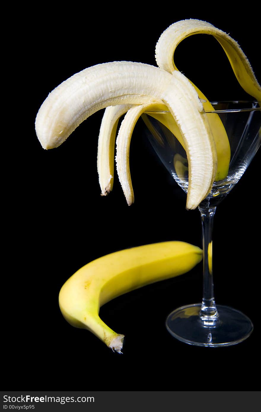 A half peeled banana presented inside a cocktail glass, isolated on a black background.  An unpeeled banana is lying on the side. A half peeled banana presented inside a cocktail glass, isolated on a black background.  An unpeeled banana is lying on the side.