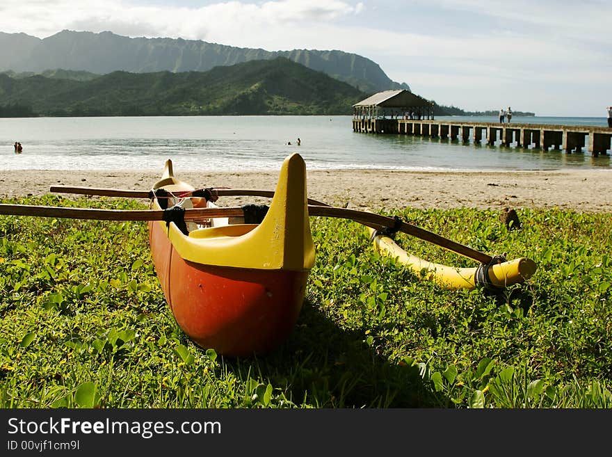 Hawaiian Outrigger Canoe on beach grass with bay, fishing pier and mountains in background.